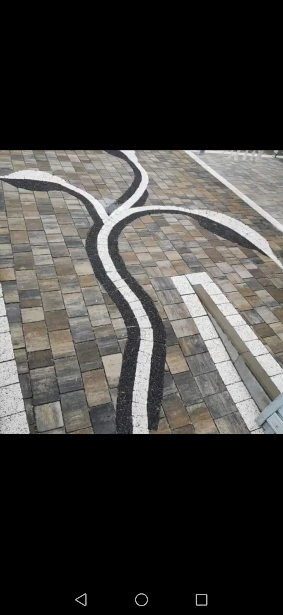 Wzór z ciemnego żwiru wkomponowany w szaro-beżową kostkę brukową, tworzący faliste ścieżki na podjeździe.