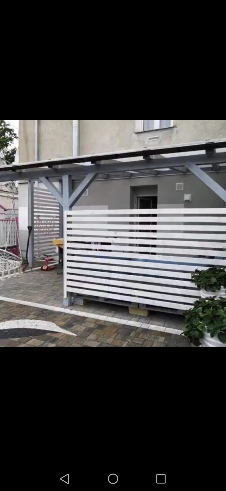 Nowoczesny taras z drewnianą pergolą i białymi panelami poziomymi, wykończony kostką brukową z dekoracyjną białą linią, donice z roślinami na tle budynku.