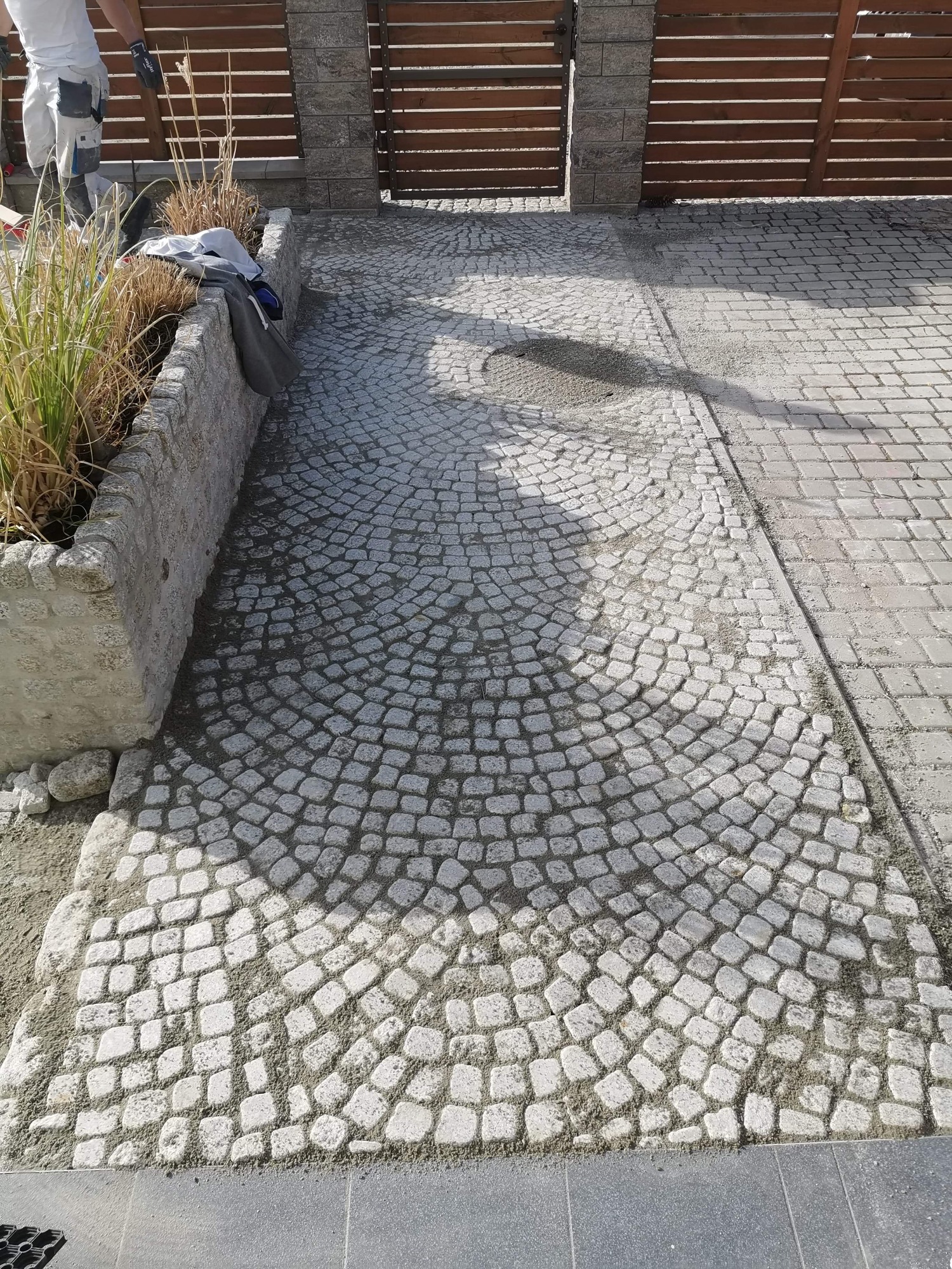 Świeżo ułożona nawierzchnia z jasnoszarej kostki granitowej, ułożonej w łukowaty wzór, z widocznym piaskiem między kostkami, przylegająca do betonowej kostki i kamiennej rabaty z suchą roślinnością...