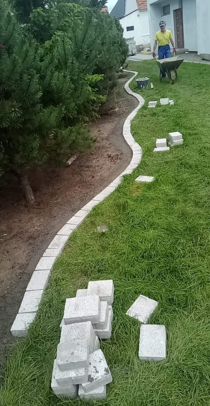 Wąska, wijąca się ścieżka z jasnoszarej kostki brukowej, częściowo ułożona wzdłuż rabaty z iglakami, z pracownikiem z taczką w tle i stertą kostki na trawniku.