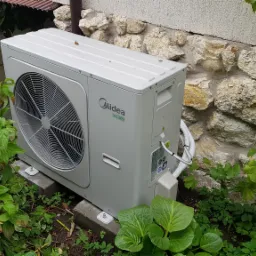 Zewnętrzna jednostka klimatyzacji Midea zamontowana na betonowych bloczkach przy kamiennej ścianie, otoczona bujną zielenią.