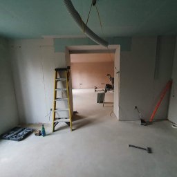 RemontujemY - Wnętrze w trakcie prac wykończeniowych: ściany obłożone płytami gipsowo-kartonowymi, widoczna drabina, narzędzia i odkurzacz przemysłowy z rurą zawieszoną u sufitu.