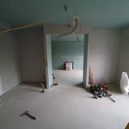 RemontujemY - Wnętrze w trakcie prac wykończeniowych: surowe ściany z płyt gipsowo-kartonowych, w tle worki z materiałem budowlanym, widoczne narzędzia i instalacja grzewcza podłogowa.