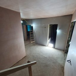 RemontujemY - Wnętrze w trakcie prac wykończeniowych: ściany pokryte gładzią, widoczna drabina, tymczasowe oświetlenie i fragment barierki, wskazujące na etap przygotowania do dalszych prac.