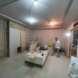 RemontujemY - Wnętrze w trakcie remontu: pracownik przechodzi obok palety płyt gipsowo-kartonowych, widoczne rusztowanie, narzędzia, worki z zaprawą, surowy mur z cegły i prowizoryczne oświetlenie.