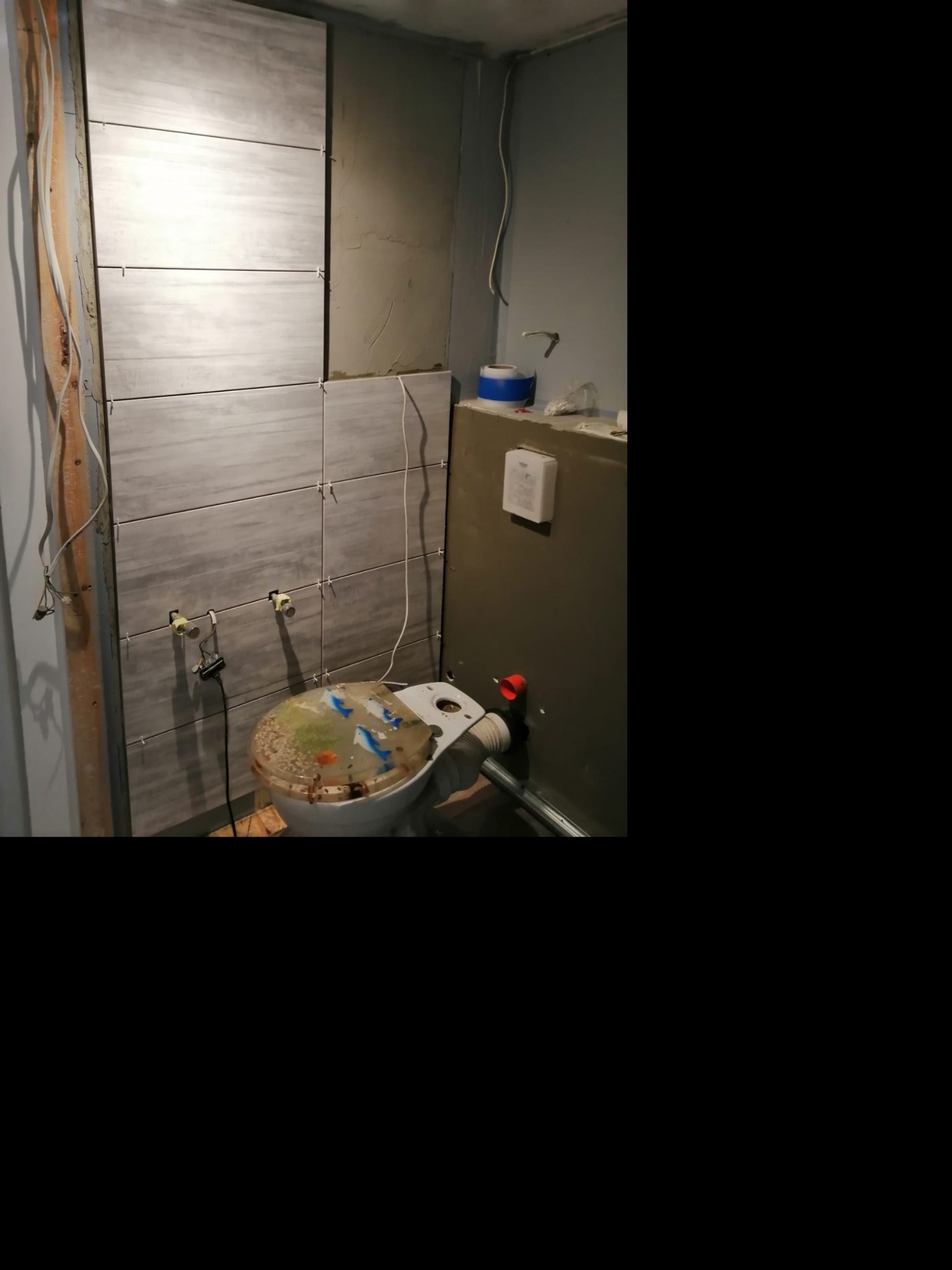 Montaż płytek ściennych w łazience, widoczne krzyżyki dystansowe, instalacja elektryczna w trakcie podłączania, toaleta z nietypową, przezroczystą deską sedesową z dekoracją wewnątrz.