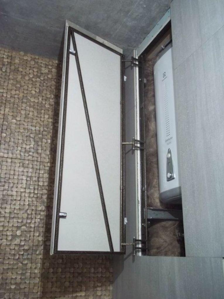 Otwarta szafka z ukrytym bojlerem w łazience, widoczne elementy konstrukcyjne i izolacja, ściana wykończona mozaiką.