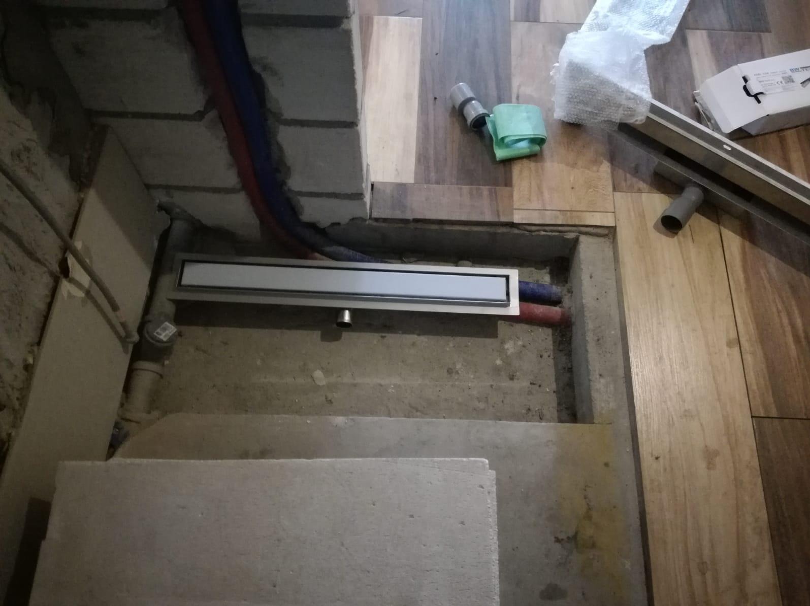 Montaż odpływu liniowego w posadzce łazienki, widoczne rury instalacji hydraulicznej i fragment ułożonej podłogi z paneli, betonowe podłoże, etap prac wykończeniowych.