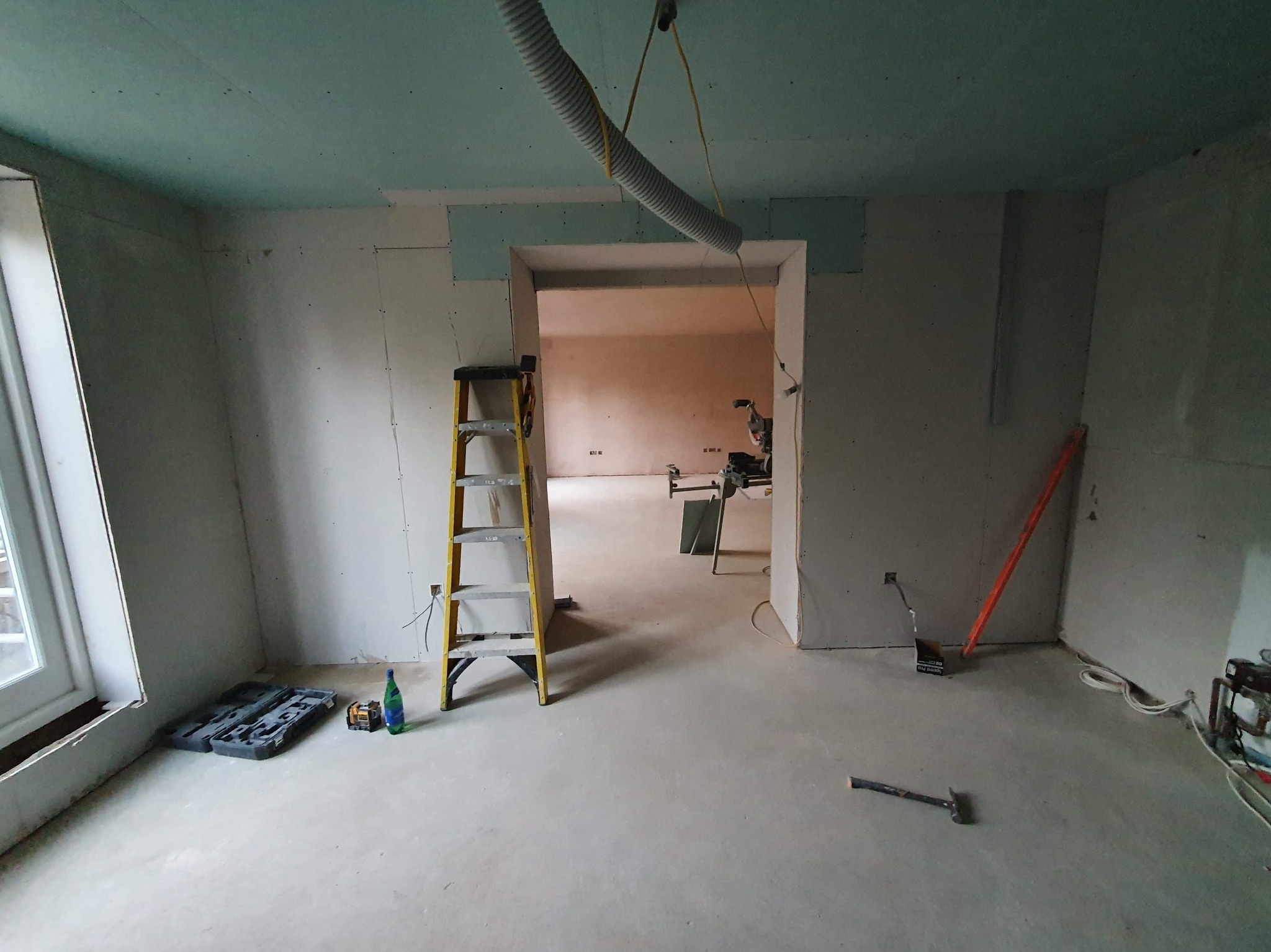 Wnętrze w trakcie prac wykończeniowych: ściany obłożone płytami gipsowo-kartonowymi, widoczna drabina, narzędzia i odkurzacz przemysłowy z rurą zawieszoną u sufitu.