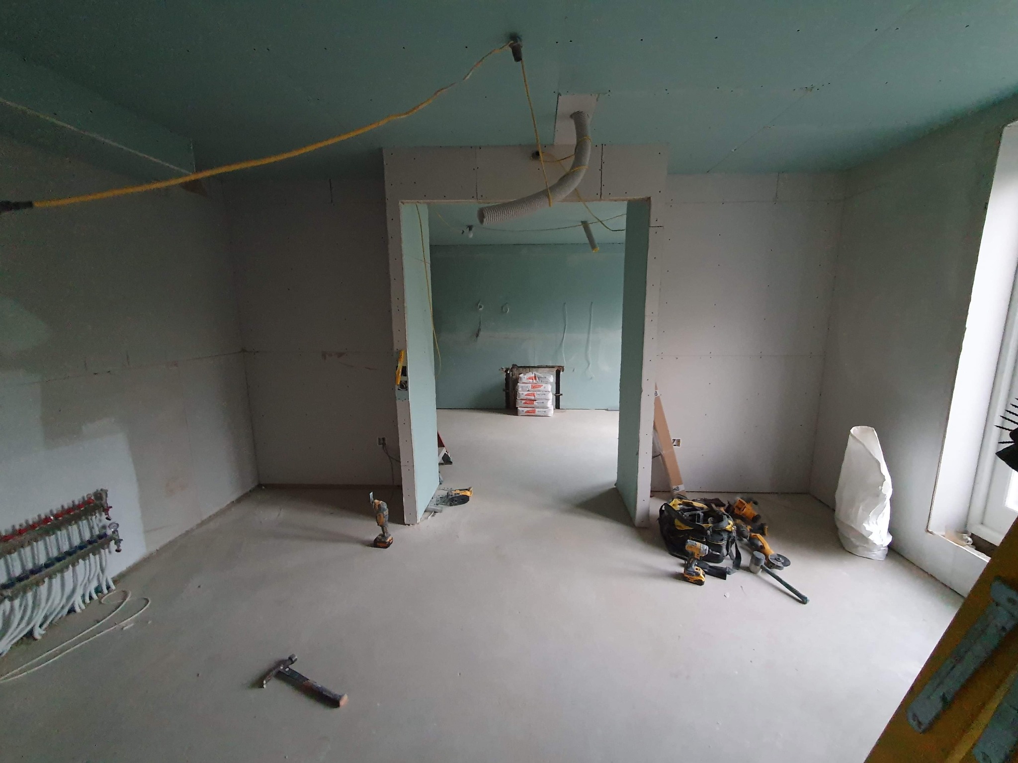 Wnętrze w trakcie prac wykończeniowych: surowe ściany z płyt gipsowo-kartonowych, w tle worki z materiałem budowlanym, widoczne narzędzia i instalacja grzewcza podłogowa.