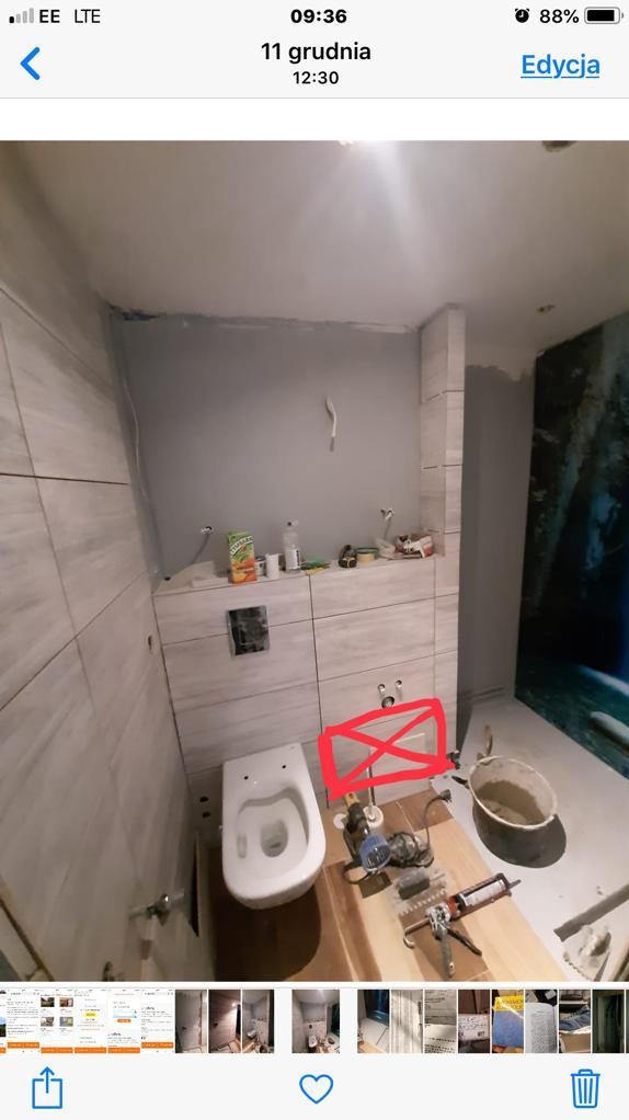 Niedokończona toaleta wisząca w trakcie montażu, widoczne narzędzia na drewnianej desce, częściowo położone płytki na ścianach, puszka soku na zabudowie stelaża.