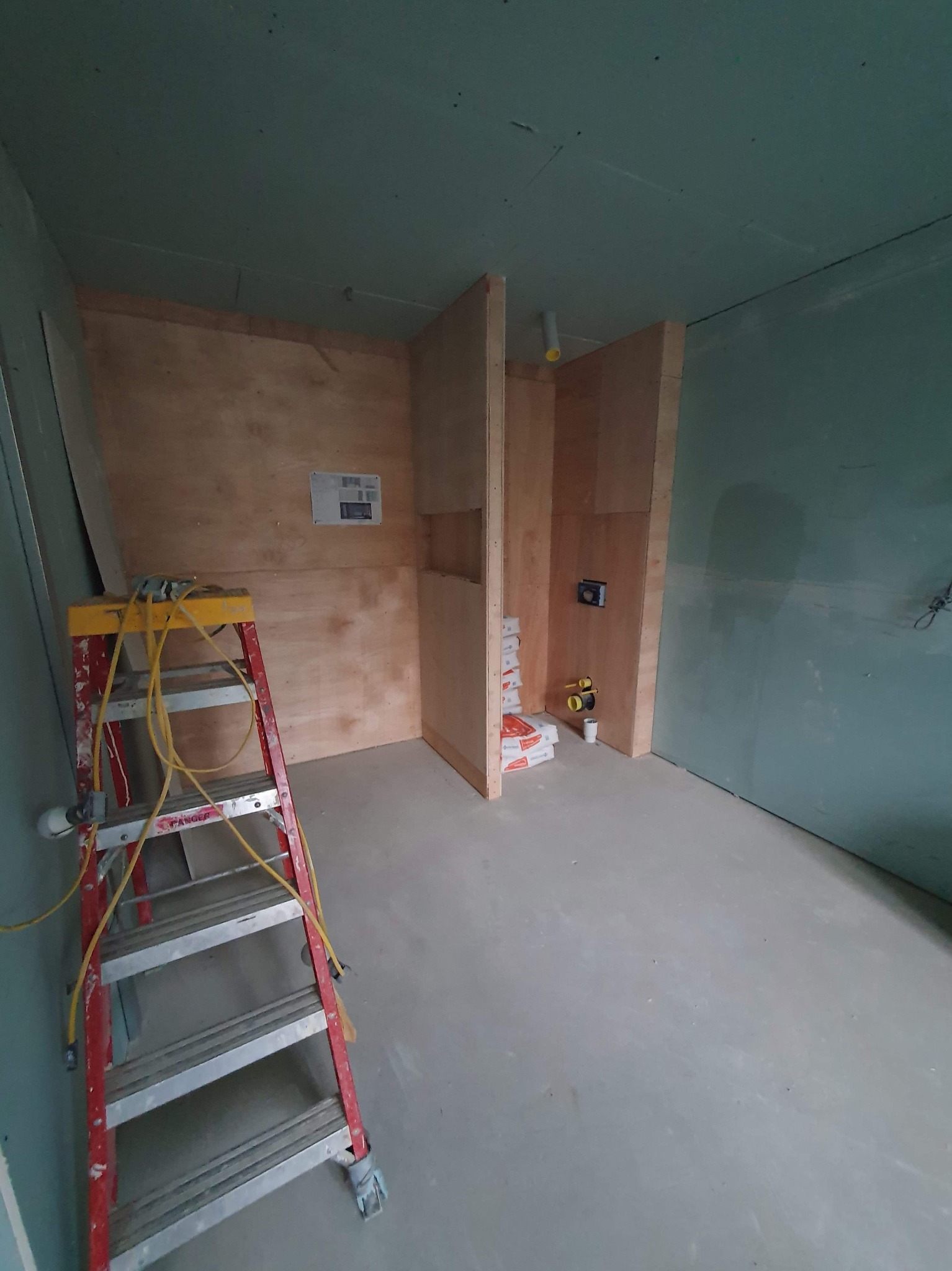 Wnętrze łazienki w trakcie prac wykończeniowych: widoczne płyty gipsowo-kartonowe na ścianach, drewniana konstrukcja zabudowy, drabina malarska z przewodami, worki z zaprawą i instalacja...