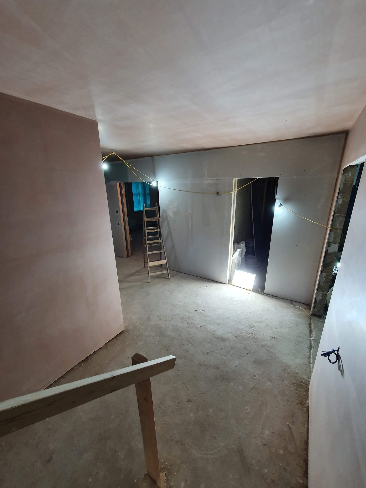 Wnętrze w trakcie prac wykończeniowych: ściany pokryte gładzią, widoczna drabina, tymczasowe oświetlenie i fragment barierki, wskazujące na etap przygotowania do dalszych prac.