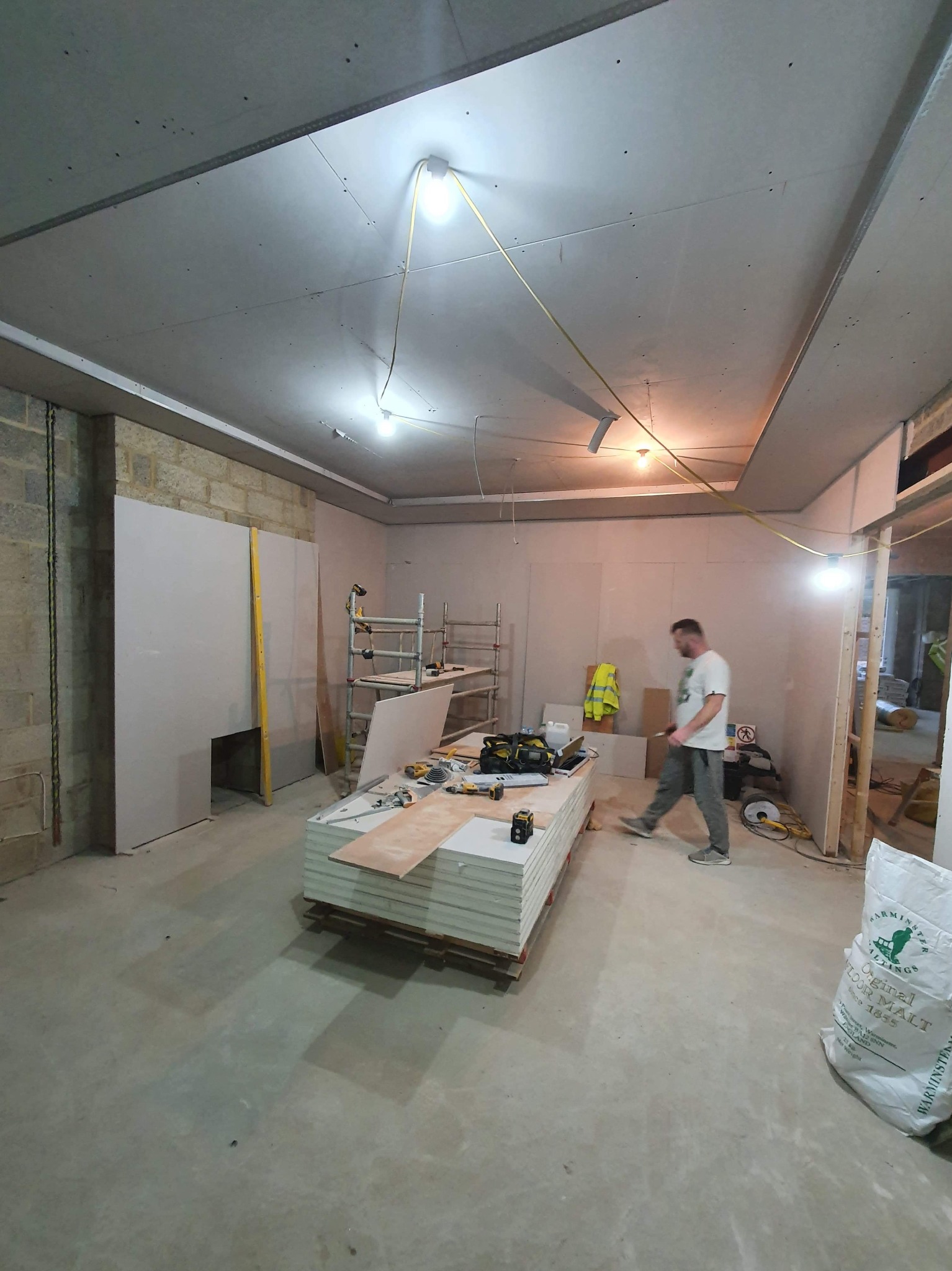 Wnętrze w trakcie remontu: pracownik przechodzi obok palety płyt gipsowo-kartonowych, widoczne rusztowanie, narzędzia, worki z zaprawą, surowy mur z cegły i prowizoryczne oświetlenie.