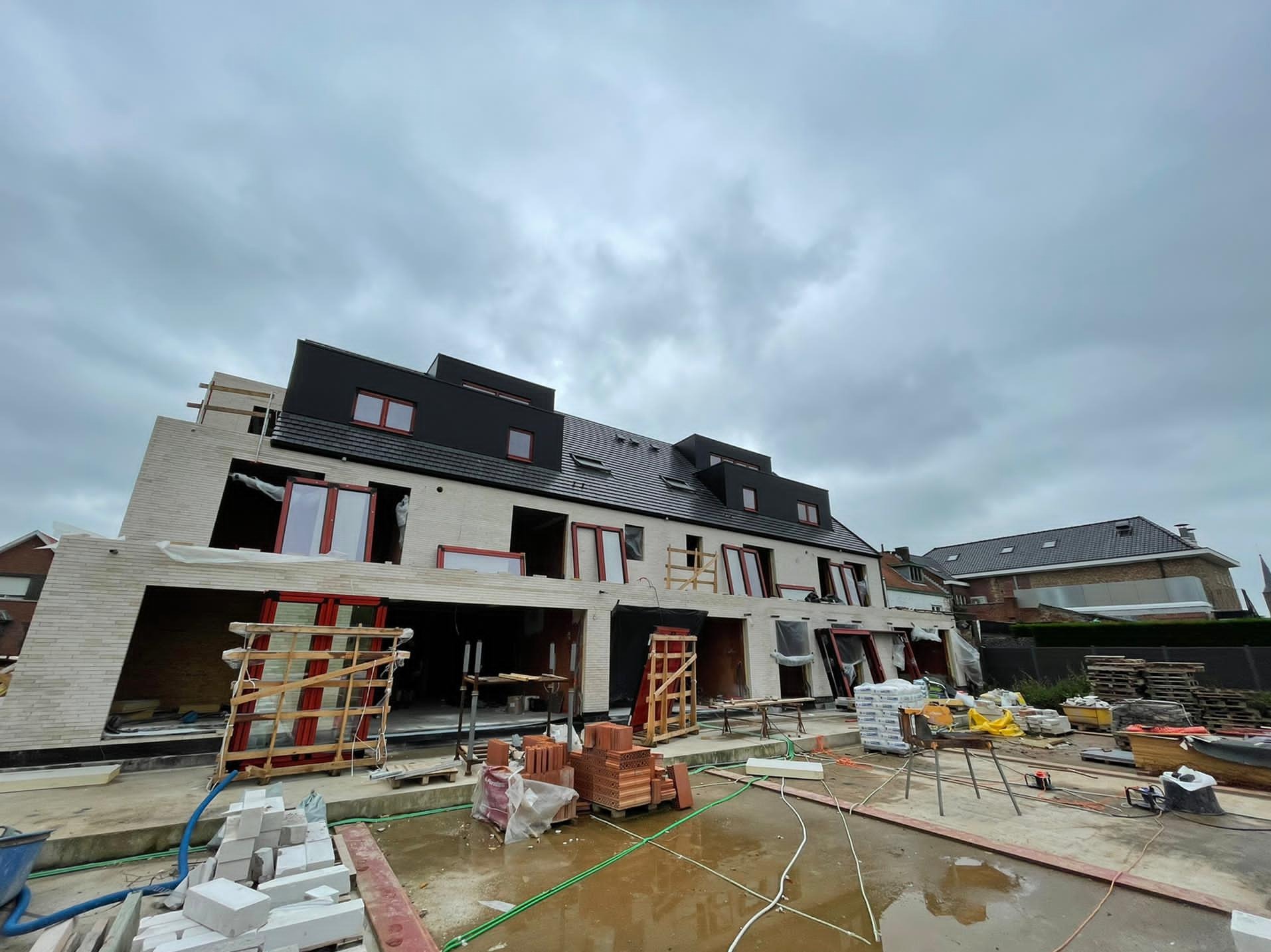 Budynek wielorodzinny w trakcie budowy: widoczne niezamontowane okna w czerwonych ramach, materiały budowlane oraz mokry plac budowy w pochmurny dzień.