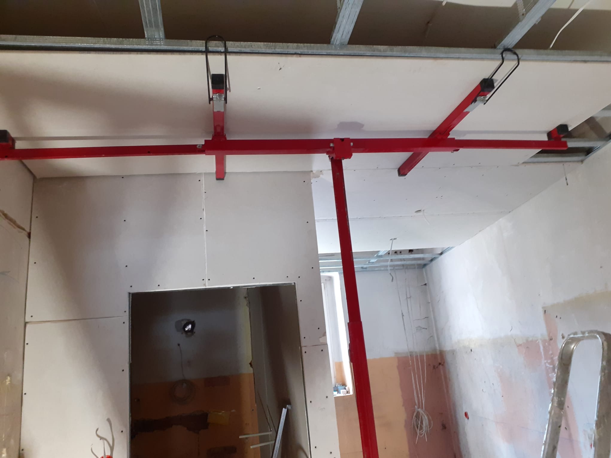 Montaż płyt gipsowo-kartonowych na suficie z użyciem czerwonego stempla montażowego, widoczne profile sufitowe i fragment ściany z widocznymi łączeniami płyt.