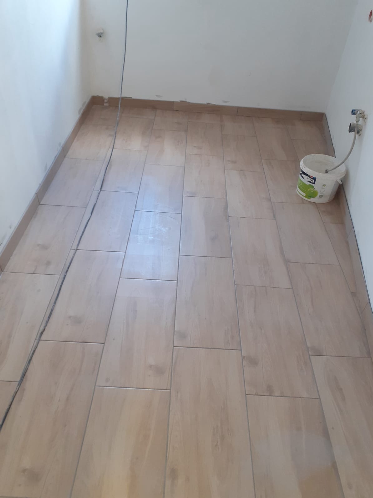 Ułożona podłoga z płytek imitujących drewno w łazience, widoczny kabel na podłodze i wiadro z farbą przy ścianie.