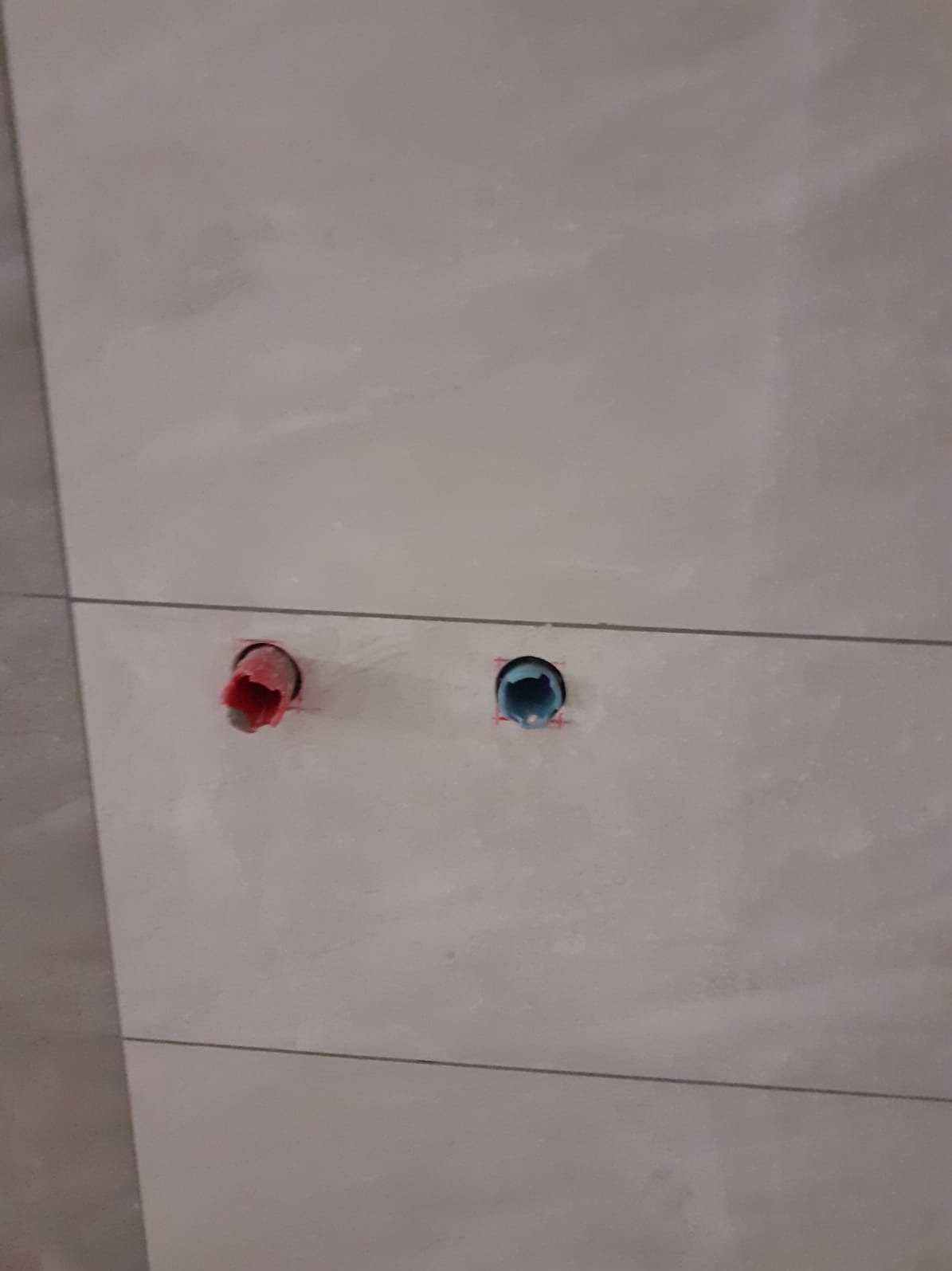 Ściana wyłożona jasnymi płytkami z widocznymi otworami instalacyjnymi z rurami w kolorze czerwonym i niebieskim.