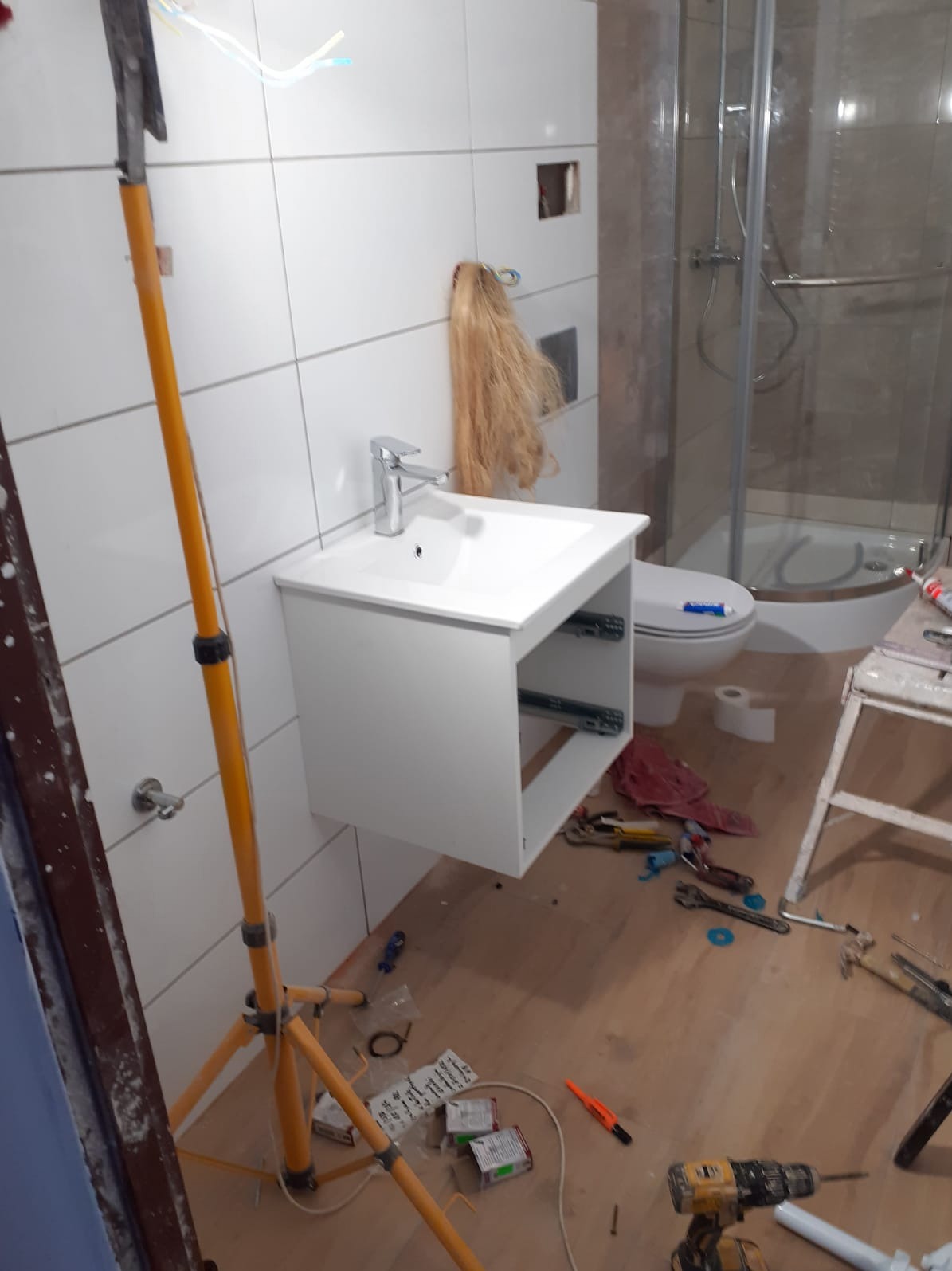 Montaż białej szafki umywalkowej z baterią w trakcie prac wykończeniowych łazienki; widoczna kabina prysznicowa, narzędzia na podłodze i lampa budowlana.