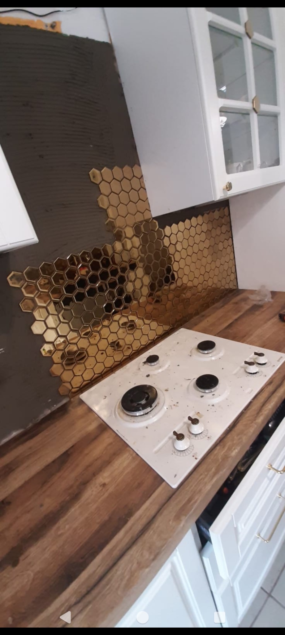 Montaż złotej mozaiki heksagonalnej na ścianie nad kuchenką gazową, widoczny klej na ścianie, drewniany blat i białe szafki kuchenne.