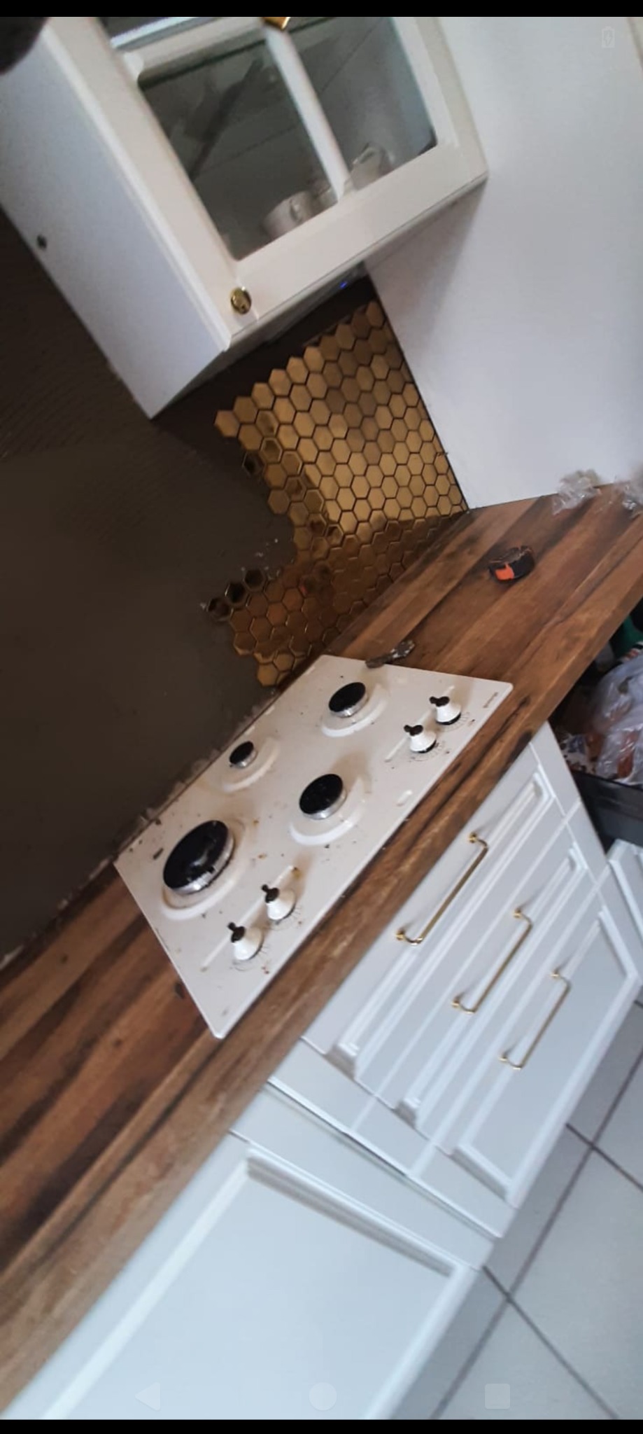 Kuchenka gazowa w trakcie montażu na drewnianym blacie, fragment ściany z mozaiką w kształcie złotych heksagonów, białe szafki z złotymi uchwytami, widoczne narzędzie miernicze na blacie.