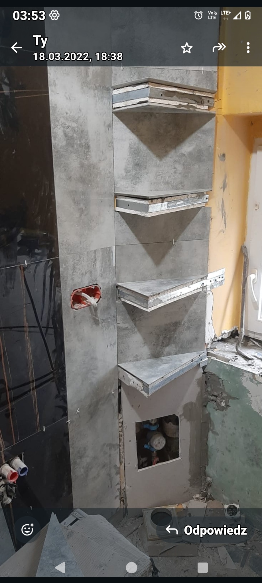 Wnęka z półkami w trakcie wykańczania w łazience, widoczne surowe płytki, fragment instalacji hydraulicznej i wystające przewody elektryczne.