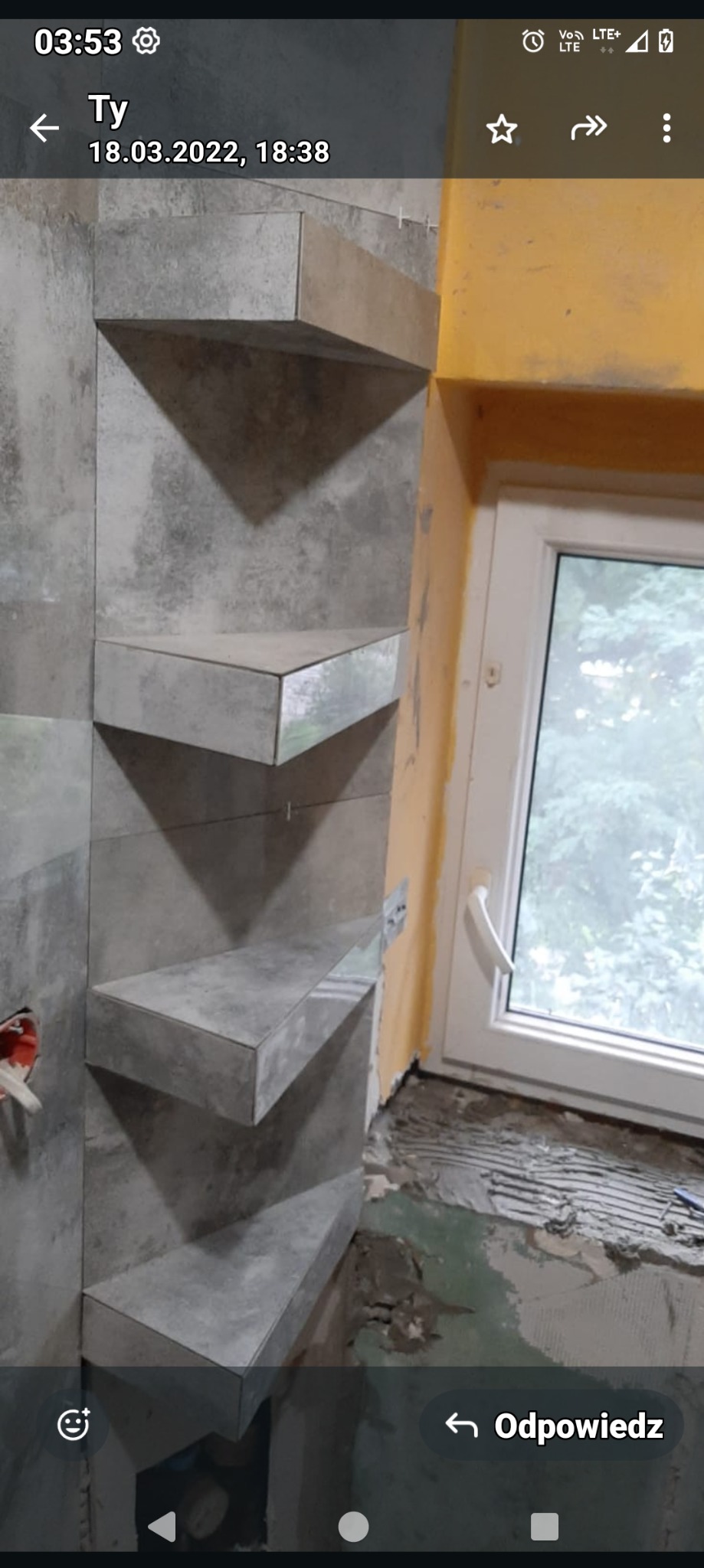 Narożna ściana w łazience z szarymi, kwadratowymi płytkami tworzącymi półki, widoczne niedokończone wykończenie przy oknie i instalacja elektryczna.