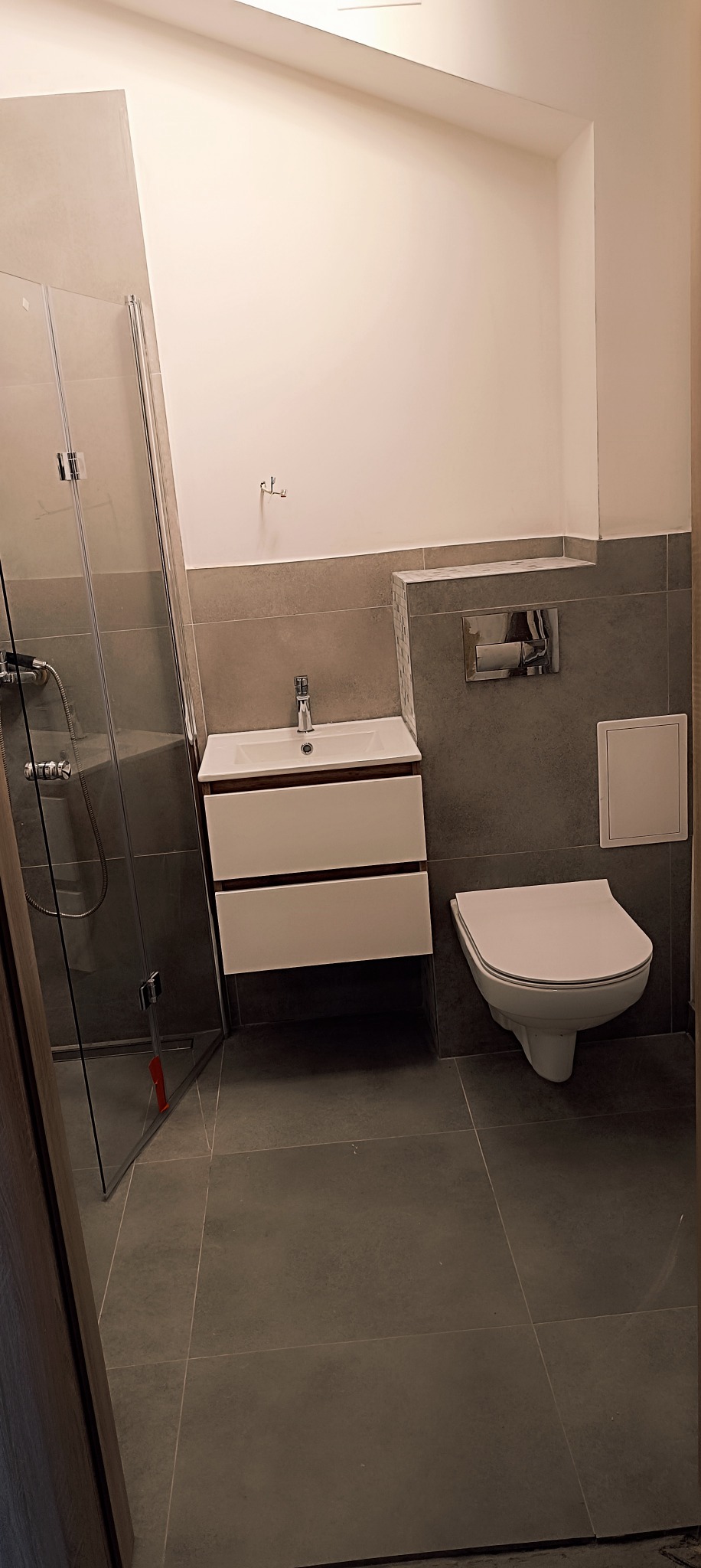 Nowoczesna łazienka po remoncie: szara glazura, biała umywalka z szafką, podwieszana toaleta i kabina prysznicowa ze szklanymi drzwiami, widoczny fragment listwy zabezpieczającej