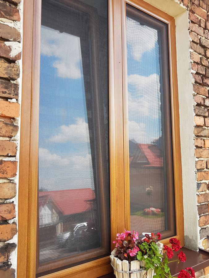 Brązowe okno z zamontowaną moskitierą ramkową, odbijające błękitne niebo z chmurami, z doniczką czerwonych kwiatów na parapecie i ceglaną ścianą w tle.