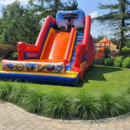 Kolorowa dmuchana zjeżdżalnia z motywem Psi Patrol na zielonym trawniku, widok z boku, w tle drzewa i budynek z pomarańczowym dachem.