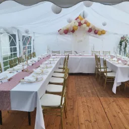 Uroczysta aranżacja wnętrza białego namiotu: długie stoły z białymi obrusami i różowymi bieżnikami, złote krzesła, dekoracje z balonów i lampionów, drewniana podłoga.