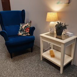 Gabinety Psychoterapii ARIADNA Katowice Opolska - Niebieski fotel uszak z poduszką w kwiaty obok białego stolika z lampką nocną i roślinką doniczkową na tle szarej ściany.