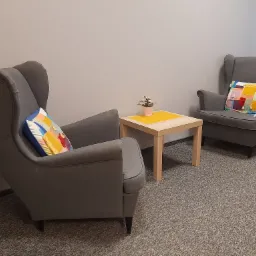 Dwa szare fotele typu uszak z kolorowymi poduszkami po bokach kwadratowego stolika z żółtym blatem i małym kwiatkiem w doniczce, na tle szarej ściany i wykładziny dywanowej.