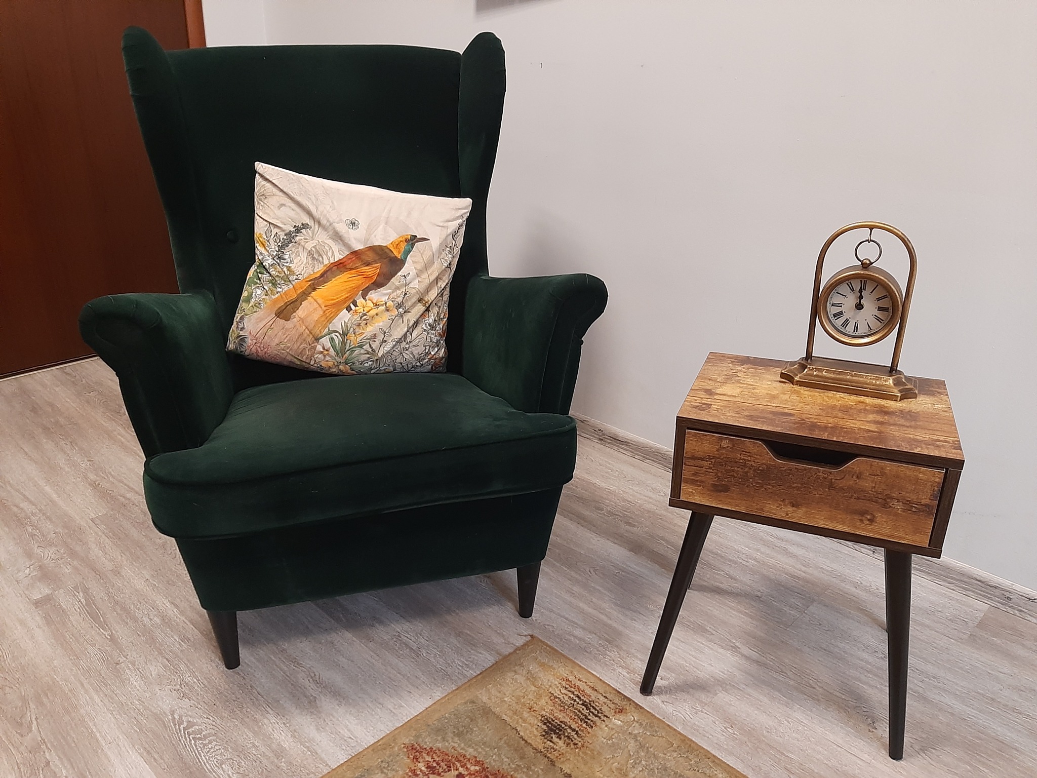 Zielony fotel uszak z poduszką z motywem ptaka, obok drewniany stolik nocny z zegarem w złotym kolorze, na jasnej podłodze.