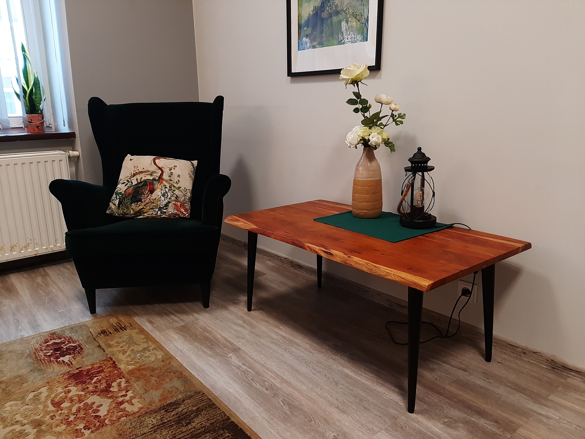 Pokój z ciemnozielonym fotelem z poduszką z motywem ptaka, drewnianym stolikiem z kwiatami w wazonie i lampą naftową, dywanem w odcieniach beżu i brązu oraz obrazem na ścianie.