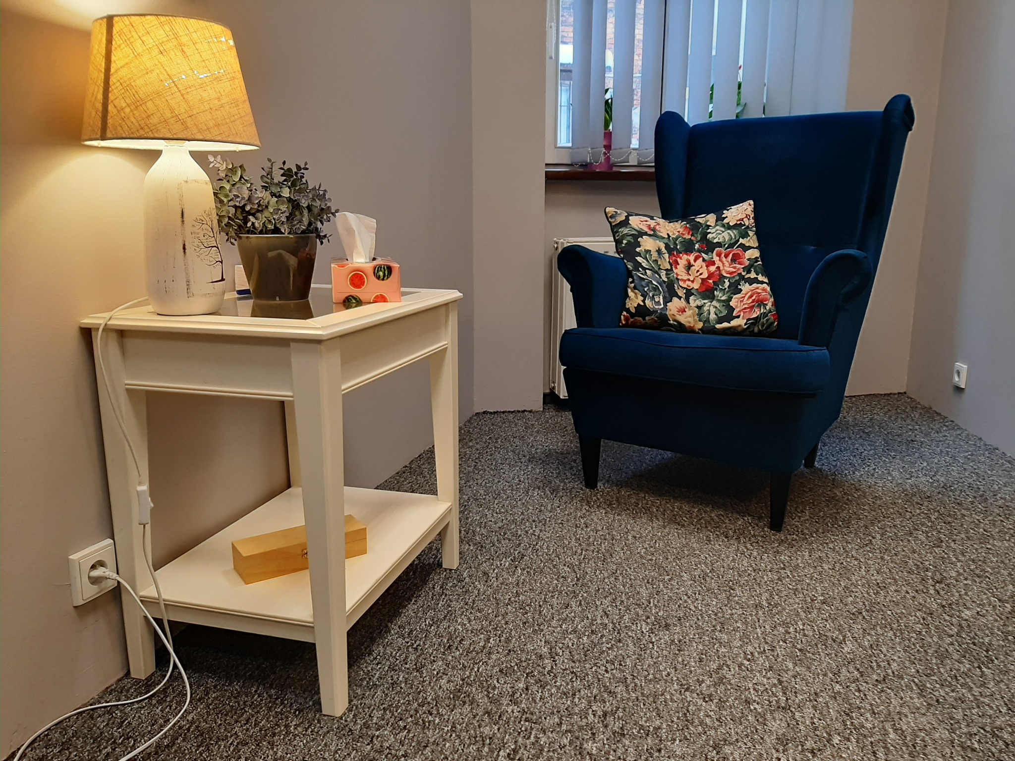Gabinet z niebieskim fotelem typu uszak, stolikiem nocnym z lampką, rośliną i pudełkiem chusteczek higienicznych.