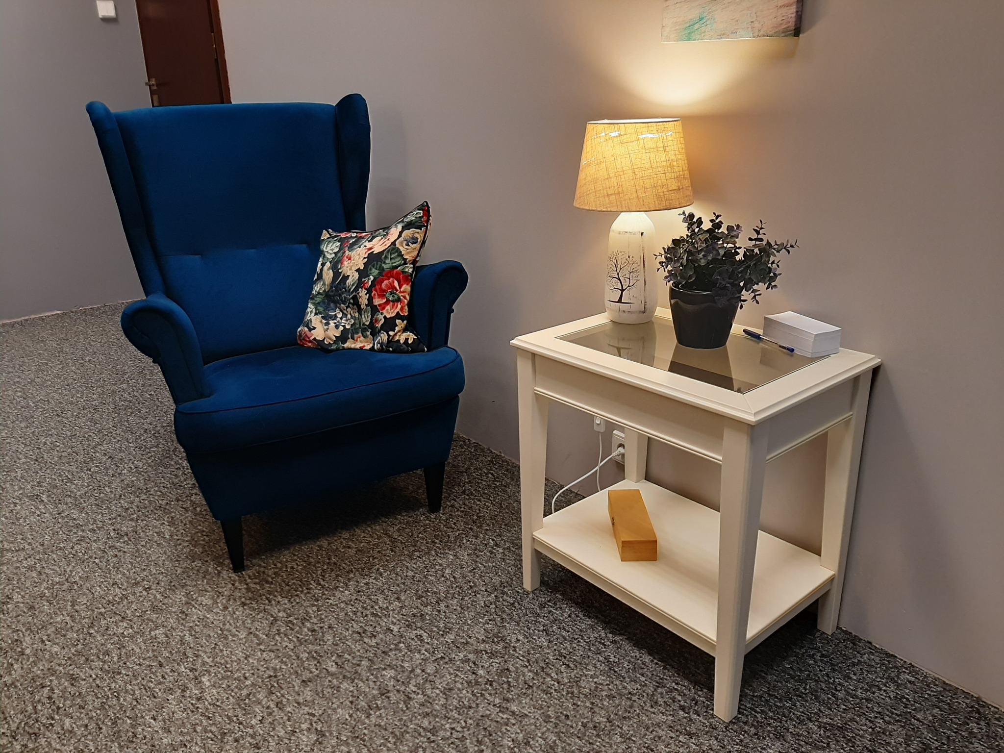 Niebieski fotel uszak z poduszką w kwiaty obok białego stolika z lampką nocną i roślinką doniczkową na tle szarej ściany.