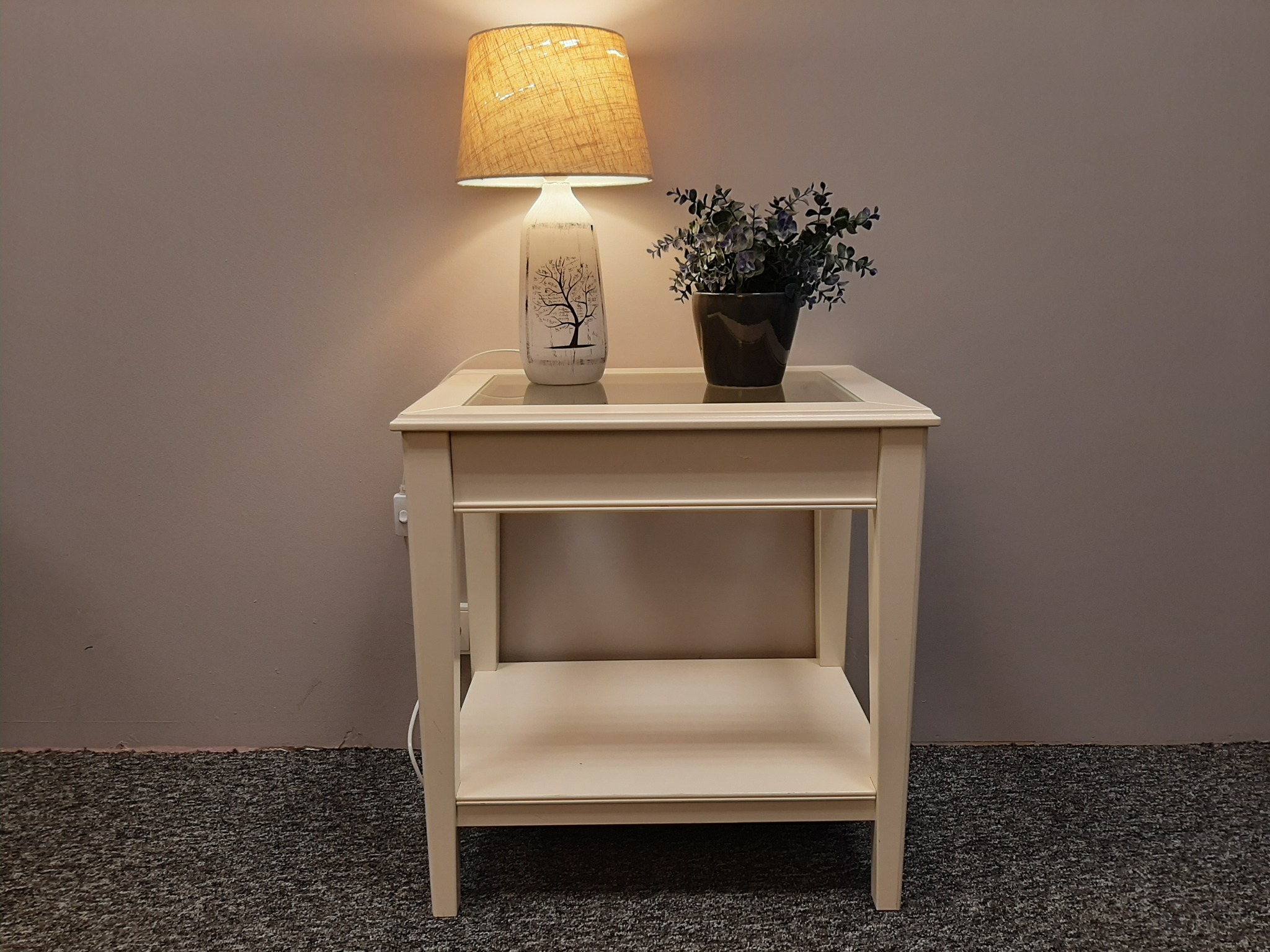 Biały stolik nocny z lampą z motywem drzewa i doniczką z rośliną na szklanym blacie, stojący na ciemnoszarym dywanie.