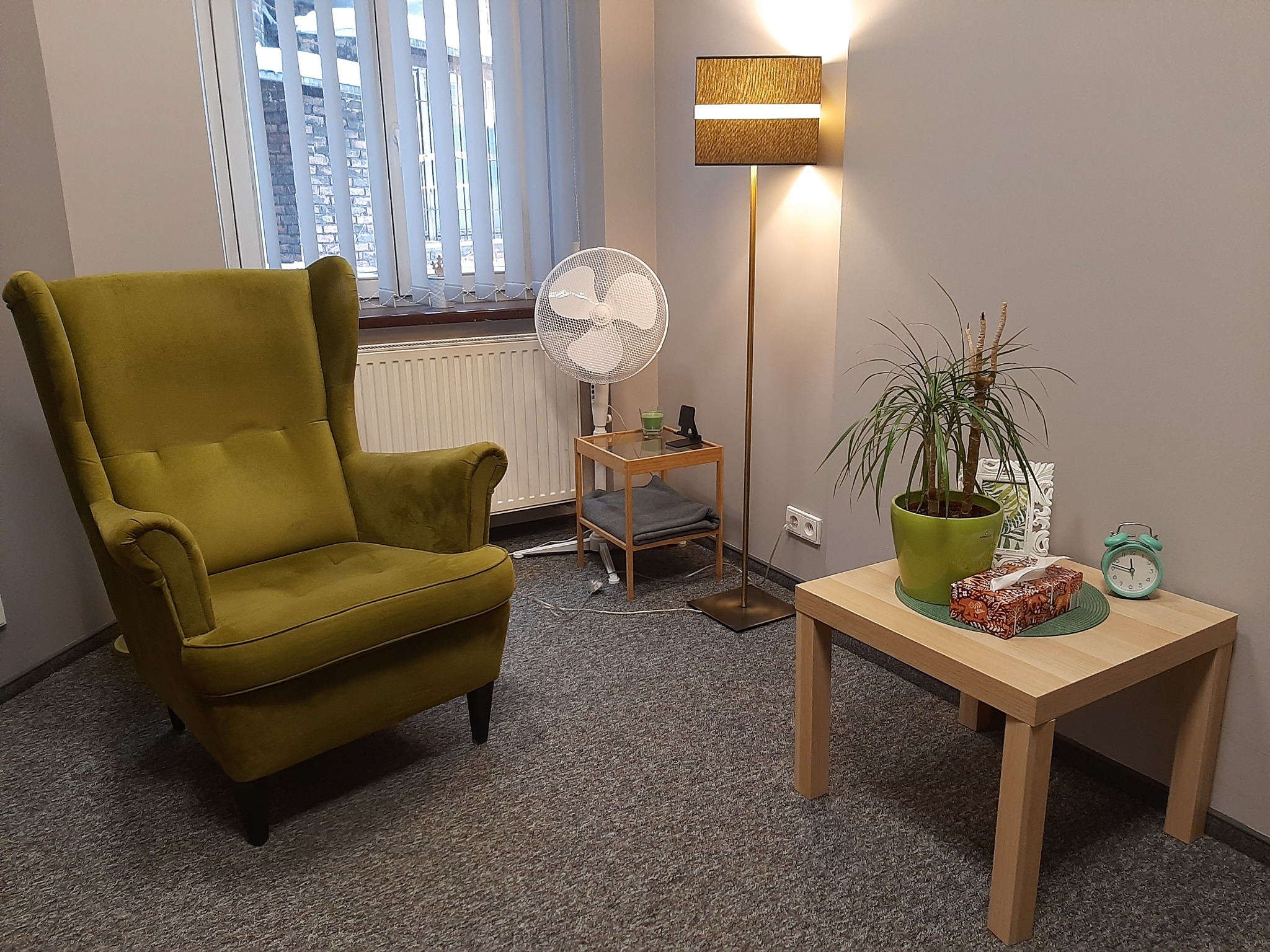 Jasne, minimalistyczne wnętrze gabinetu z zielonym fotelem uszakiem, stolikiem z rośliną doniczkową i chusteczkami, wentylatorem oraz lampą podłogową z abażurem w odcieniach brązu.