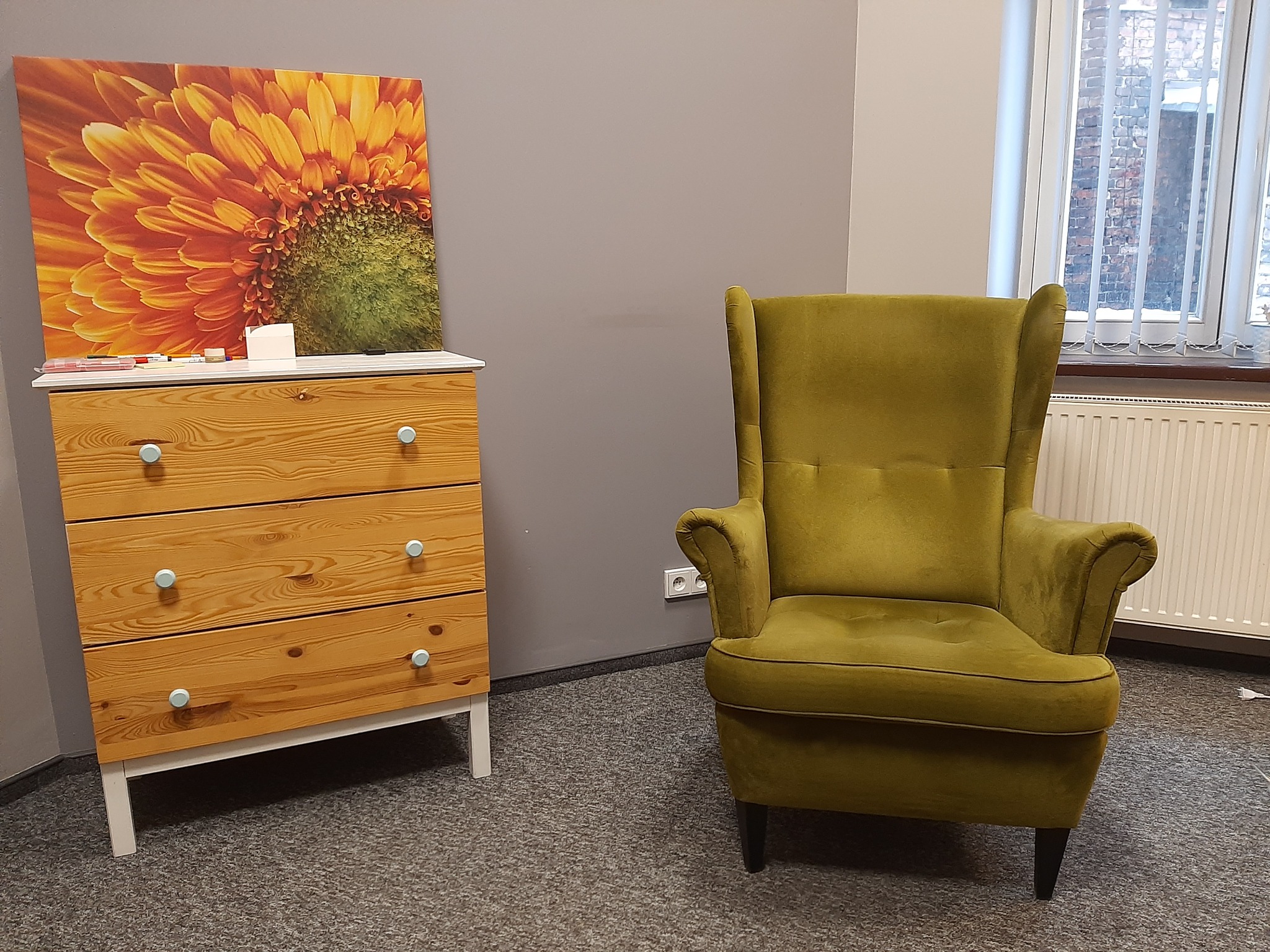 Pokój z komodą z obrazem słonecznika i zielonym fotelem typu uszak, okno z żaluzjami w tle.