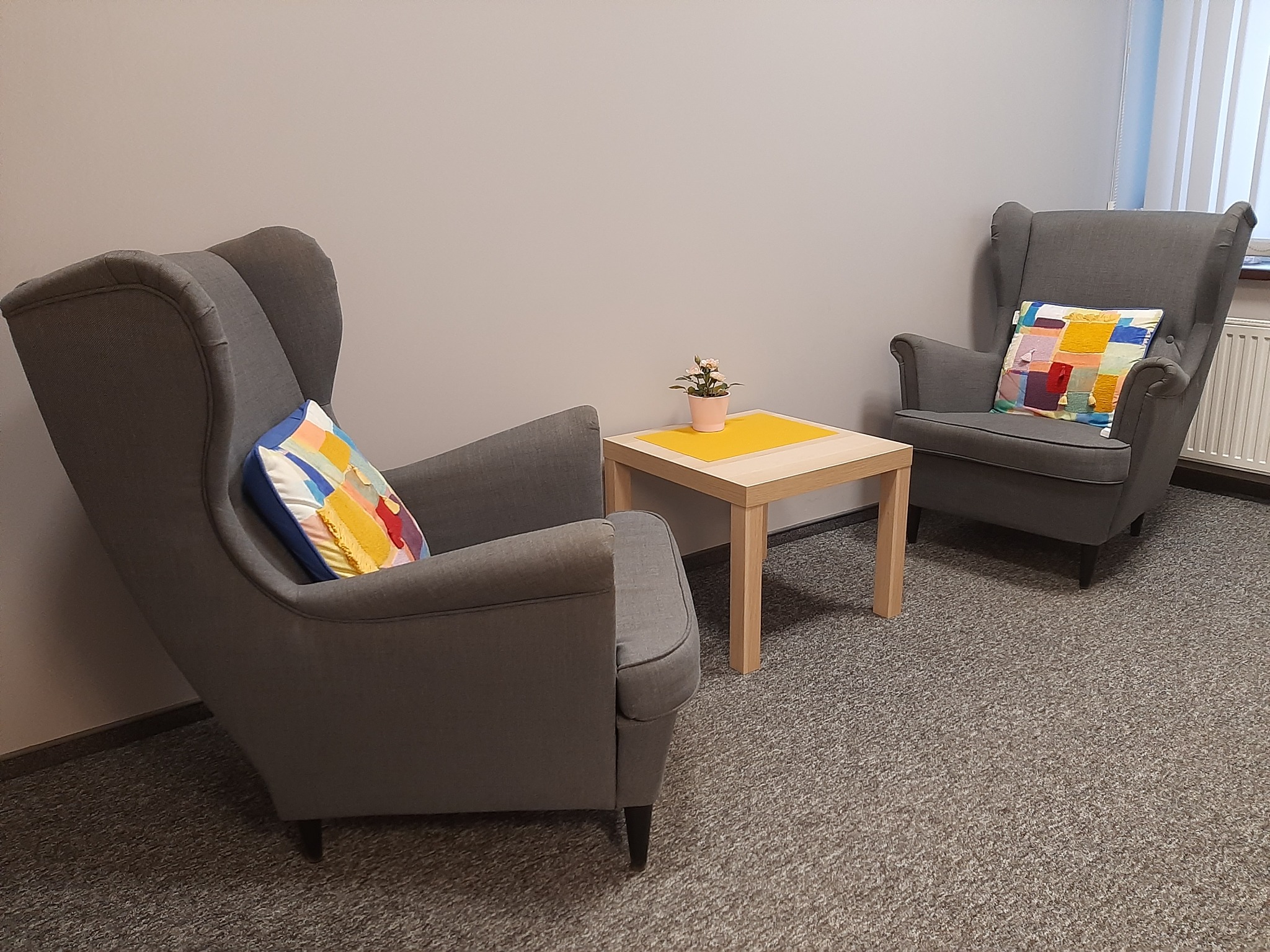 Dwa szare fotele typu uszak z kolorowymi poduszkami po bokach kwadratowego stolika z żółtym blatem i małym kwiatkiem w doniczce, na tle szarej ściany i wykładziny dywanowej.