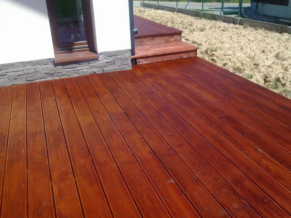 Ciemnobrązowy taras drewniany z widocznymi schodami, przylegający do białego budynku z szarą podmurówką z kamienia dekoracyjnego.