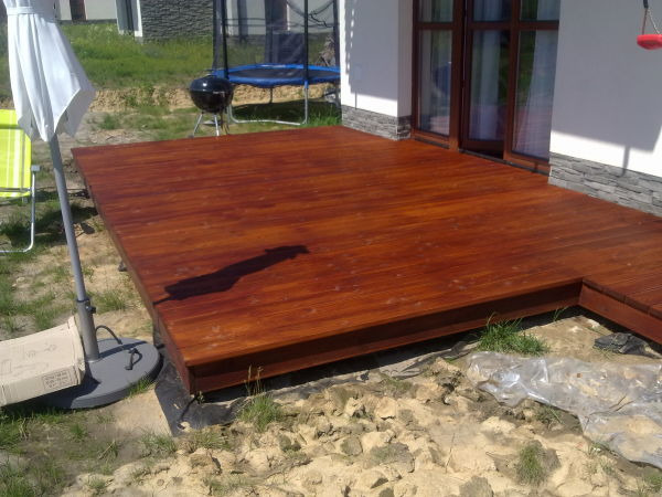 Świeżo zaimpregnowany, brązowy podest z desek przy budynku z oknami i szarym cokołem, widoczne trawnik, parasol i trampolina w tle.