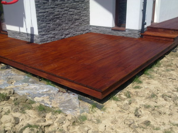 Nowoczesny, brązowy podest z drewna przy domu z kamienną elewacją, z widocznymi schodkami i folią ochronną na ziemi.