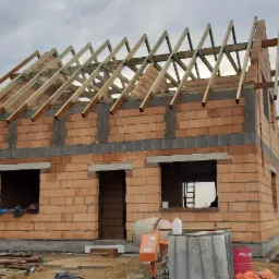 Surowy stan domu z czerwonej cegły z częściowo ukończoną konstrukcją dachu z drewnianych belek, widoczna betoniarka i worki z materiałami budowlanymi na placu budowy.