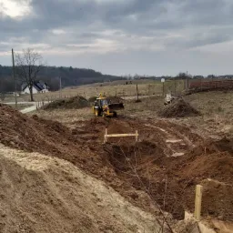 Żółta koparko-ładowarka w trakcie prac ziemnych na nierównym terenie, widoczne wykopane rowy i pryzma ziemi, w tle dom i zalesione wzgórza pod pochmurnym niebem.