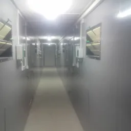 Długi, sterylny korytarz z szarymi ścianami, oświetlony lampami sufitowymi, z widocznymi otworami wentylacyjnymi z regulowanymi klapami i przewodami elektrycznymi.