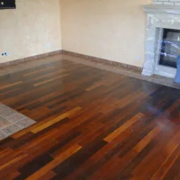 Błyszcząca, ciemna podłoga z desek ułożonych w jodełkę, z obramowaniem z płytek ceramicznych, częściowo zakryta folią ochronną przy kominku.