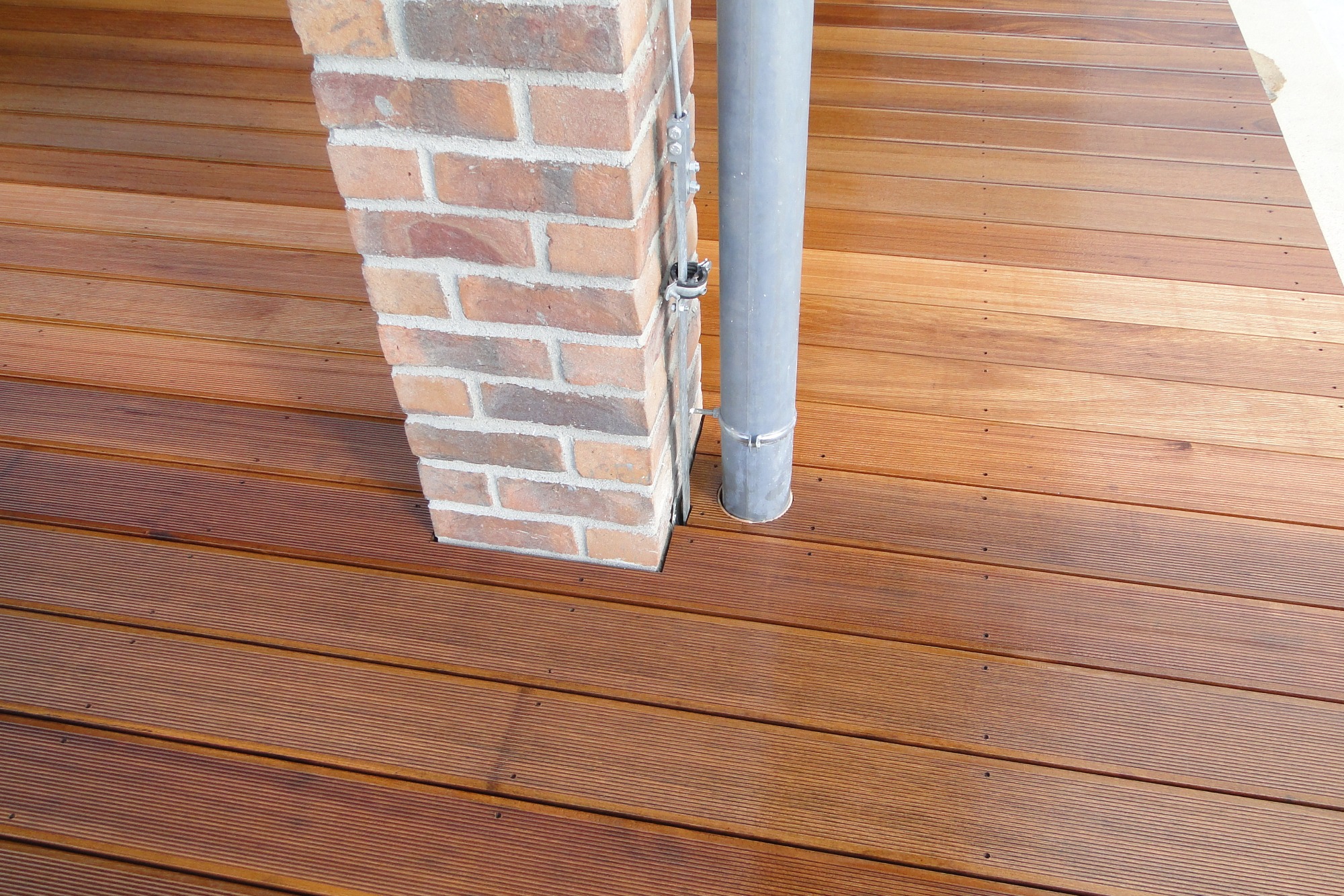 Ułożona deska tarasowa z drewna egzotycznego, przylegająca do ceglanego filaru i rury spustowej