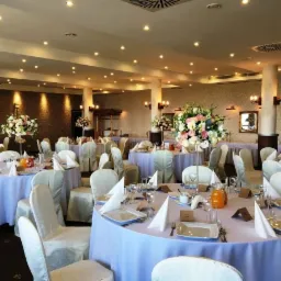 Uroczysta sala balowa z okrągłymi stołami nakrytymi jasnofioletowymi obrusami, białymi krzesłami i kompozycjami kwiatowymi na stołach, przygotowana na przyjęcie.